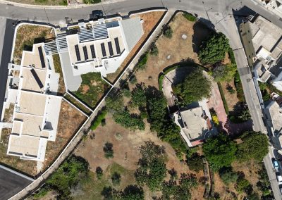 Vista aerea di due edifici bianchi con tetti piani e pannelli solari, circondati da giardini e da un’area residenziale con alberi e strade.